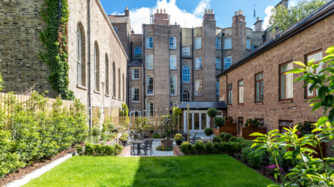 Spacious garden courtyard with lush greenery, outdoor seating, and historic brick architecture under a bright blue sky.