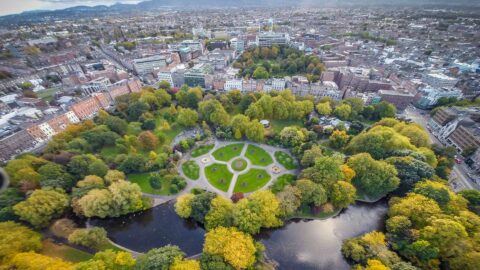 Aerial view of lush city park with walking paths, vibrant trees, and a serene pond, surrounded by charming urban landscape.