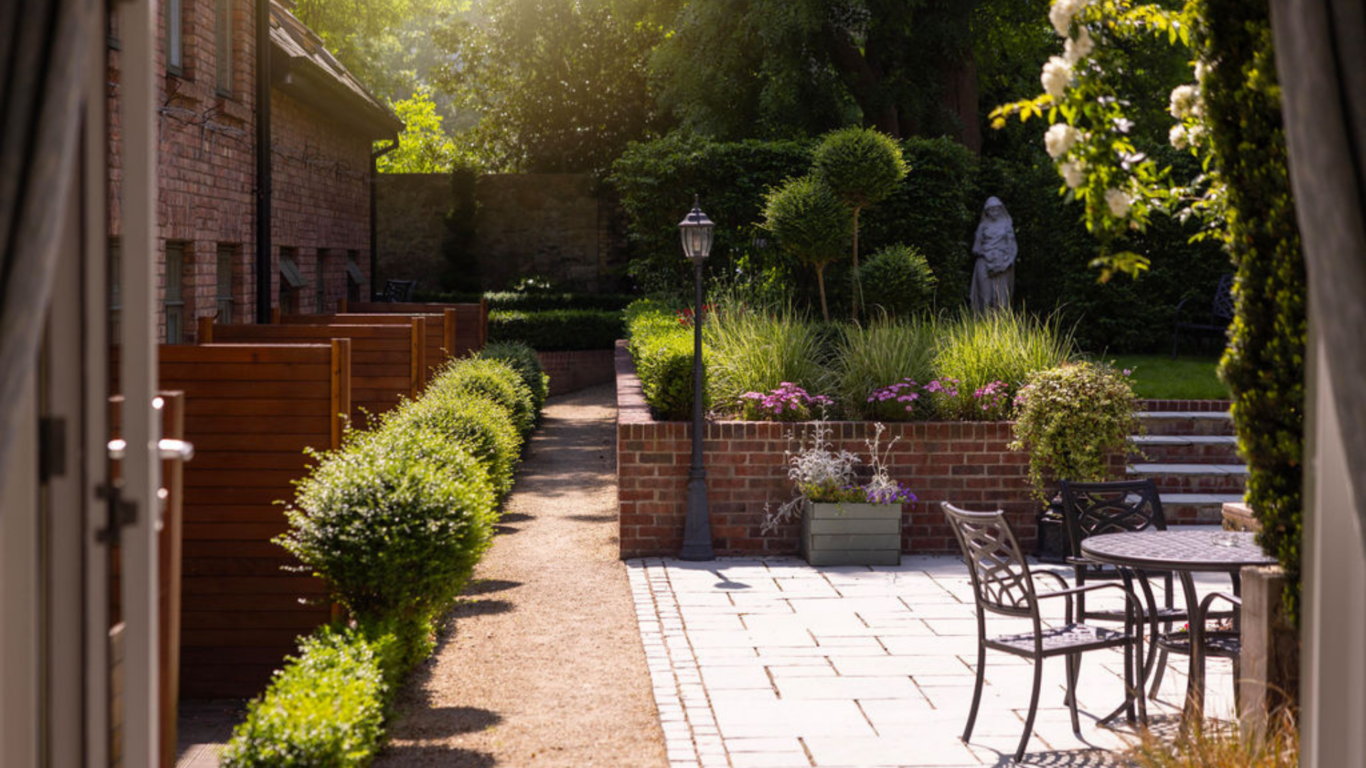 Charming hotel garden with sunlit patio, elegant iron chairs, manicured bushes, and a serene statue amidst vibrant flowers.