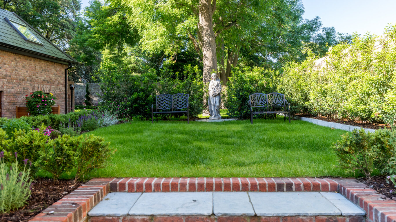 Relax in serene hotel garden with lush greenery, charming benches, and stone statue beneath shady trees.